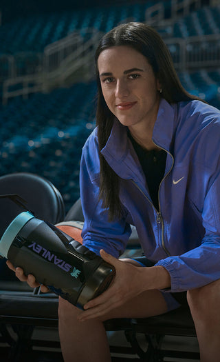 Caitlin Clark holding Stanley water jug sitting on bench chairs in basketball arena