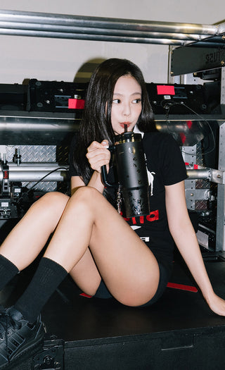 Jennie in a black graphic t-shirt holding a black Stanley tumbler inside an industrial-style setting.