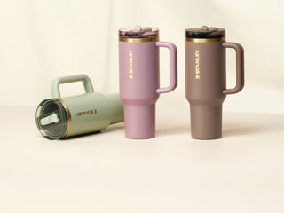 Three Stanley tumblers in sage green, pink, and brown with gold accents displayed on a neutral surface.
